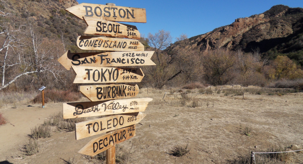 A list of different destinations on a wooden sign at a crossroads, mountains and arid land sit in the background